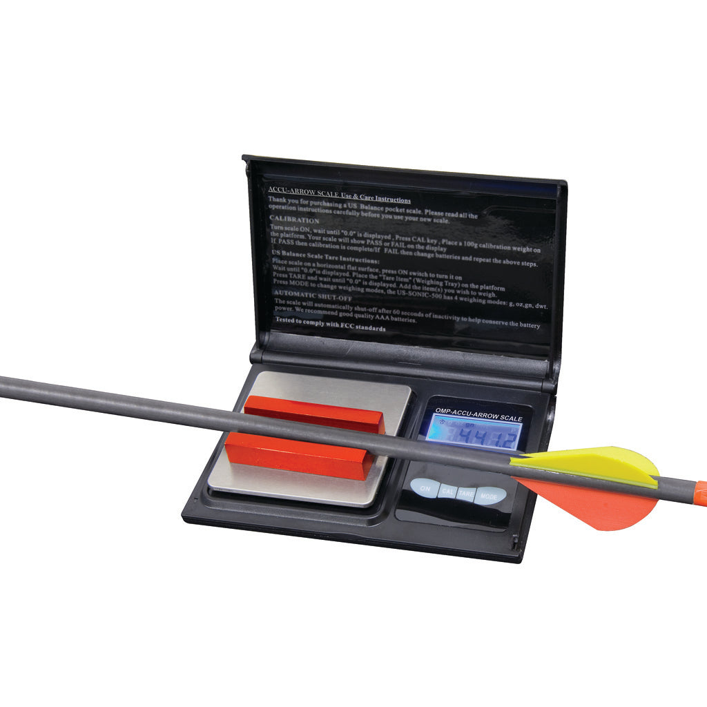 Open black digital grain scale with a lit display and two red calibration weights, a fletched arrow lying across the case, on a white background.