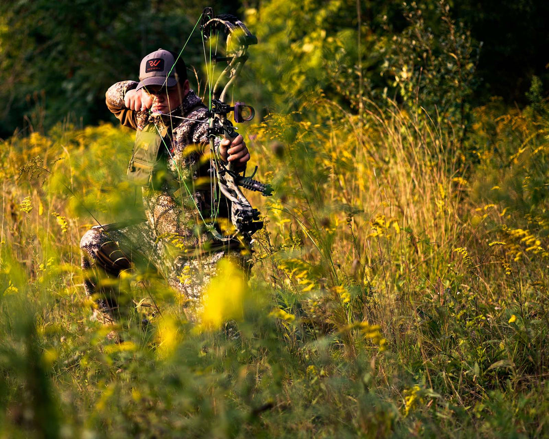 Bowhunter at full draw at golden hour in a field of tall goldenrod and grass, wearing camouflage and an orange-logo cap, aiming a compound bow.