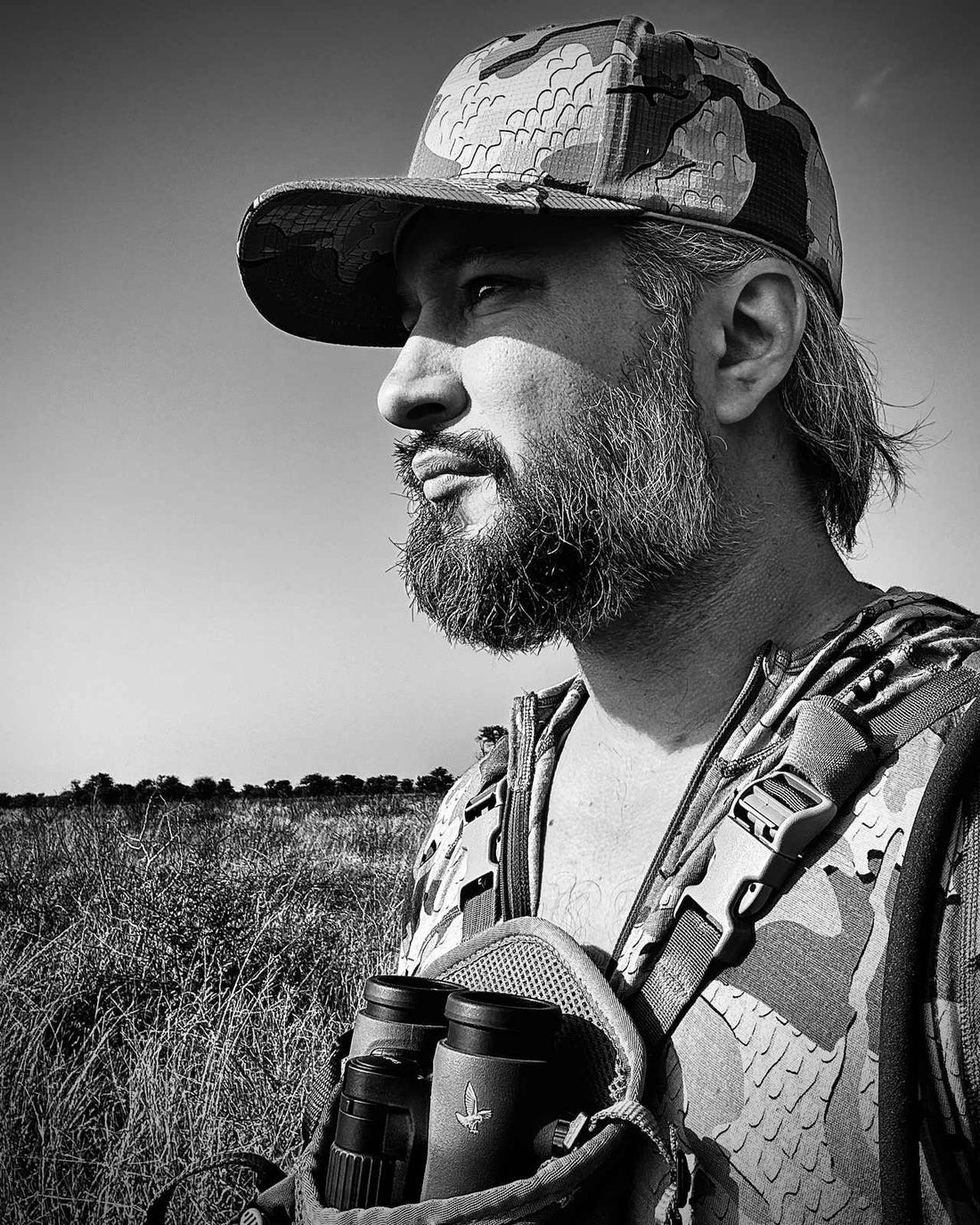 Black and white portrait of a bearded hunter in a camouflage cap with binoculars around his neck, looking out over open grassland.